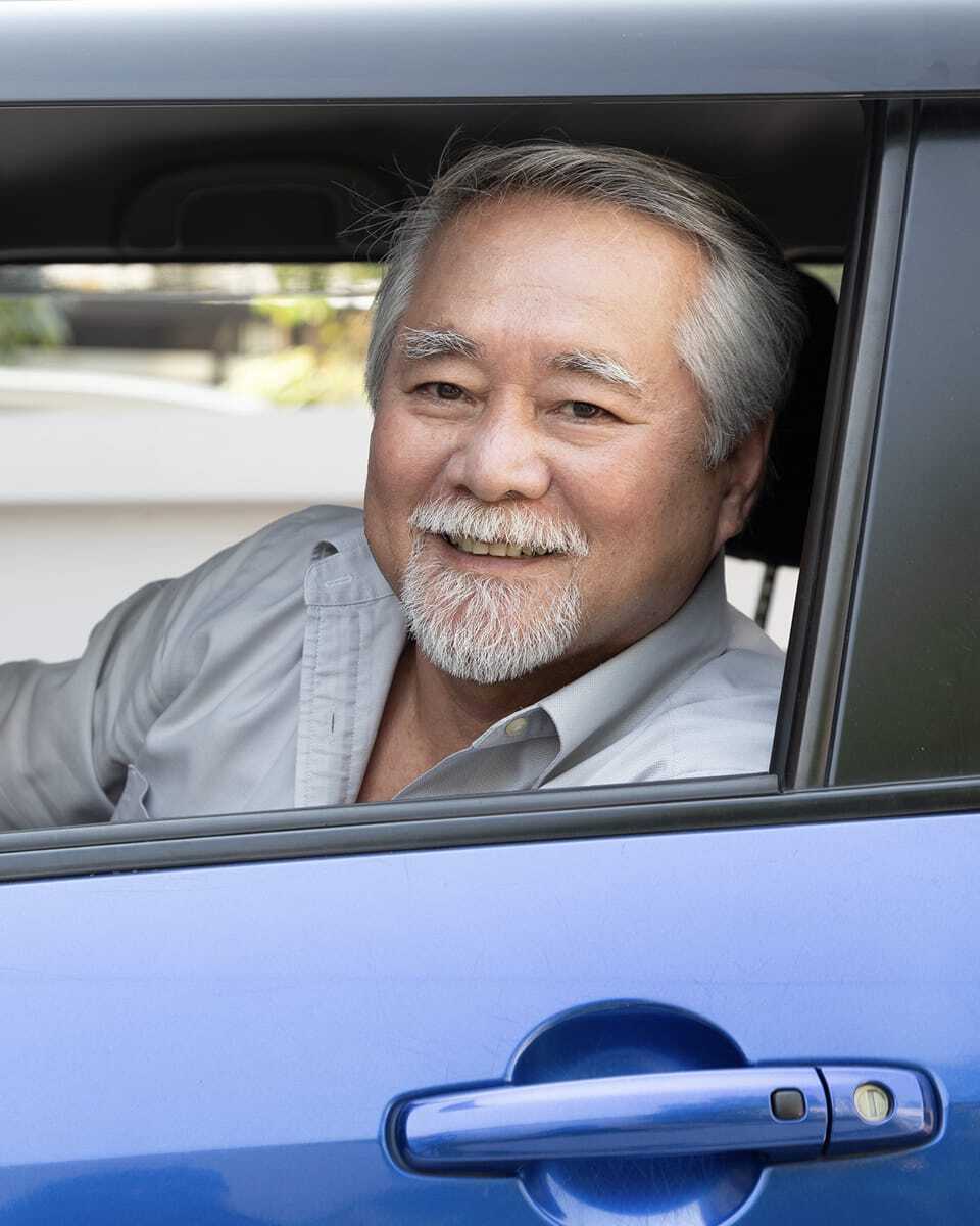 Asian man sitting in a car looking at us through the driver's side window.